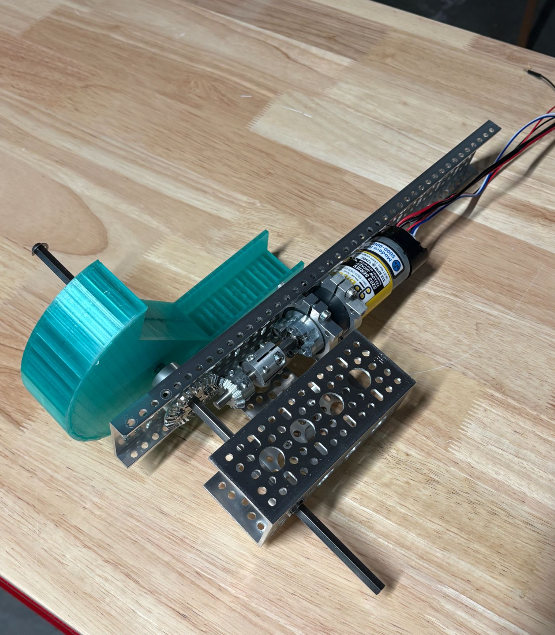 Photo of the first topsoil scoop prototype on a test bench: a green 3D-printed scoop on a shaft, driven by a geared DC motor mounted on an aluminum channel, resting on a wooden table.