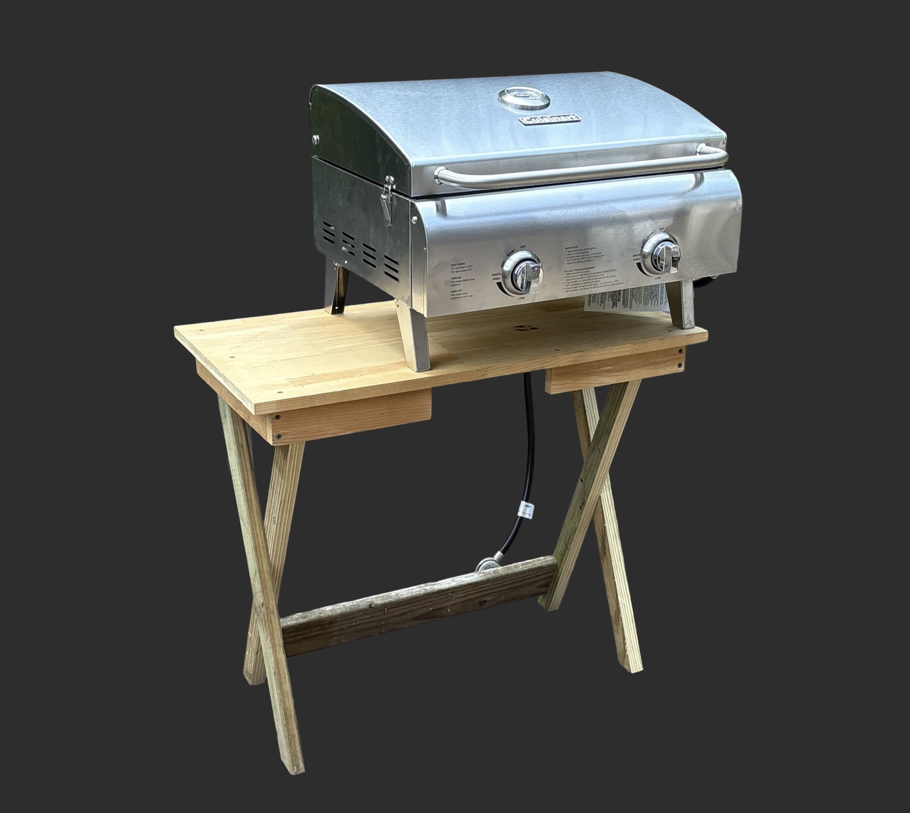 Grill table made from scrap wood: a tabletop on crossed, X-shaped legs joined by a lower rail, holding a portable stainless-steel propane grill.