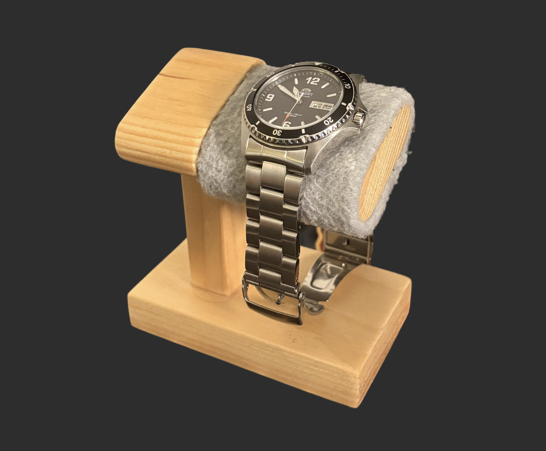Watch stand made from scrap wood: a rounded wooden post on a rectangular base, with a gray fleece-covered cushion holding a steel dive watch.